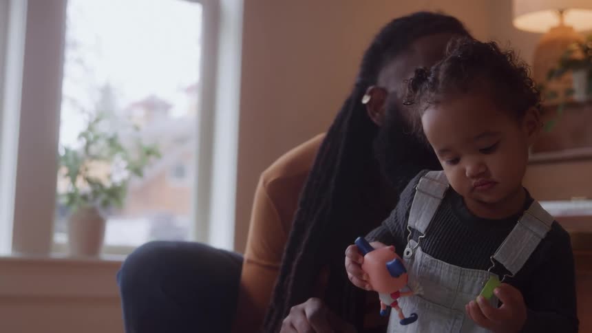 Child plays with toys while an adult watches smiling in the background