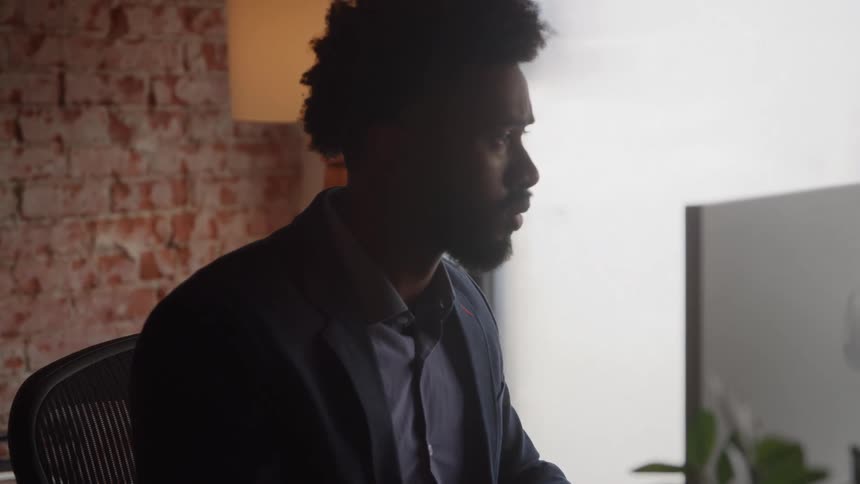 Young black man wearing a blue suit sitting down and typing on a laptop computer indoors during day time