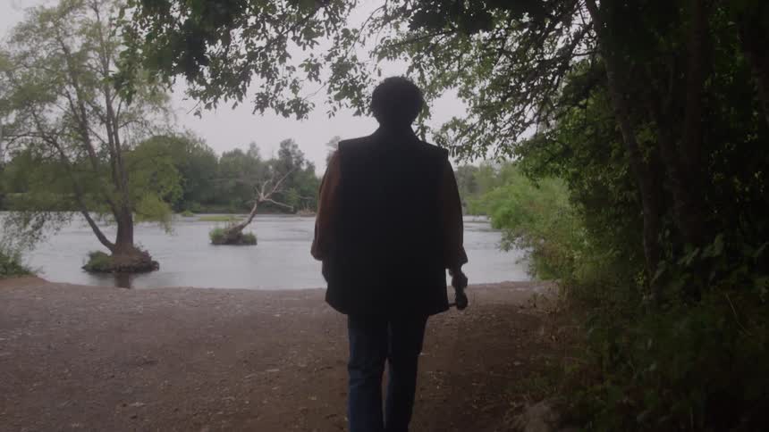A dark silhouette of a person wearing a vest and jeans is walking toward a wide river or lake while holding a fishing rod on a cloudy day framed by dark green foliage