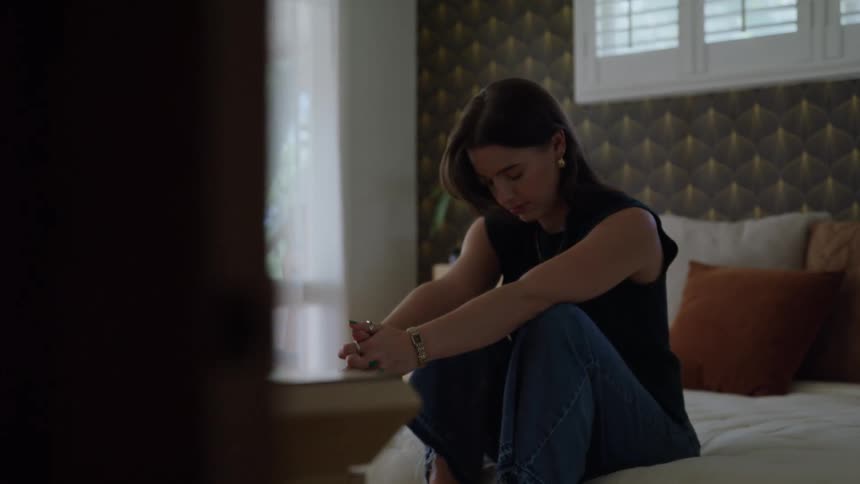 A young woman sits on a bed at midday her gaze cast downward in quiet sadness hands gently extended in front of her