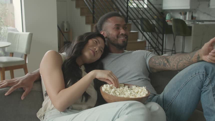 The video shows a man and a woman sitting on a couch in a modern brightly lit living room sharing a bowl of popcorn they are both smiling and looking offcamera suggesting they are enjoying watching something together