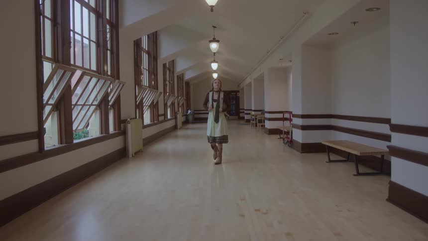 The video shows a woman with blonde hair wearing a scarf and a lightcolored dress as she walks down a long welllit hallway the hallway features tall windows with open panes along one side and benches against the opposite wall