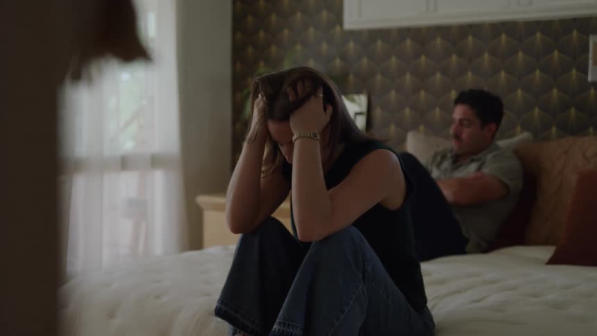 A woman sits hunched over on a bed with her knees pulled up looking distressed as she holds her head in her hands a man is seated further back on the bed with his arms crossed appearing angry and distant from her