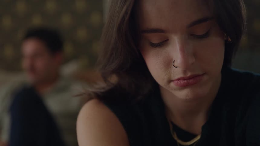 A dark close view shows a pensive woman with dark hair and a nose ring closing her eyes wearing a black shirt and gold necklace with a blurred man sitting in the dark background