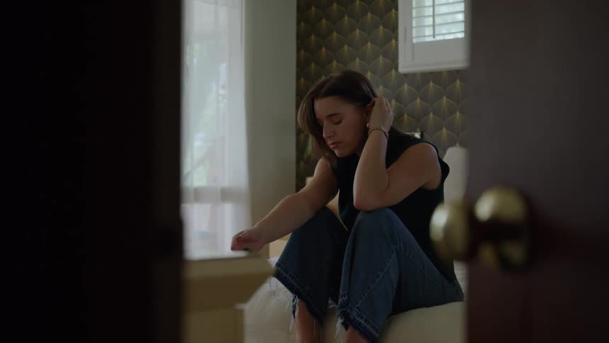 A view through an open doorway shows a woman sitting slouched on a bed the woman holds her head and then rests her hands on her folded legs the bedroom features patterned wallpaper and a window with white shutters