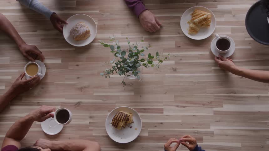 The video provides a highangle overhead shot of a group of people gathered around a light woodgrain tableseveral hands enter the frame placing cups of coffee or tea and plates with pastries and toast around a centerpiece of leafy green sprigs sug