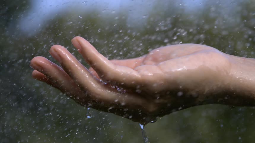 The video is a closeup of an open hand holding its palm up as water or rain falls down the scene is outdoors with a blurred background and droplets collect and stream off the fingers
