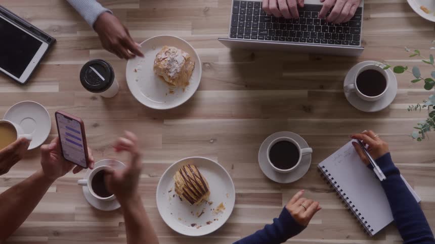 Small group of people typing on devices and writing on notebooks while eating pastries and drinking coffee indoors during day time