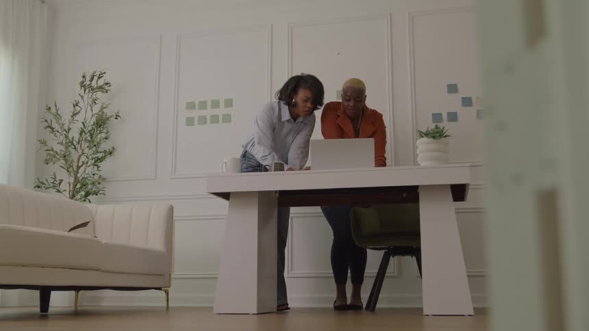 Woman stands at a table with a laptop computer as another woman stands talking and pointing at the screen