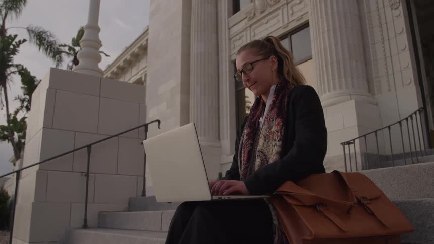 A young woman wearing glasses sits on an outdoor staircase at sunset typing on a laptop placed on her lap with a bag beside her