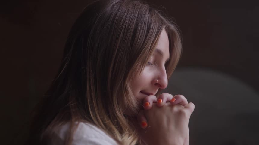 A woman with her eyes closed and head bowed her hands are clasped together near her chin her hair falls forward around her face the scene is dimly lit with the woman partially illuminated by a soft light source