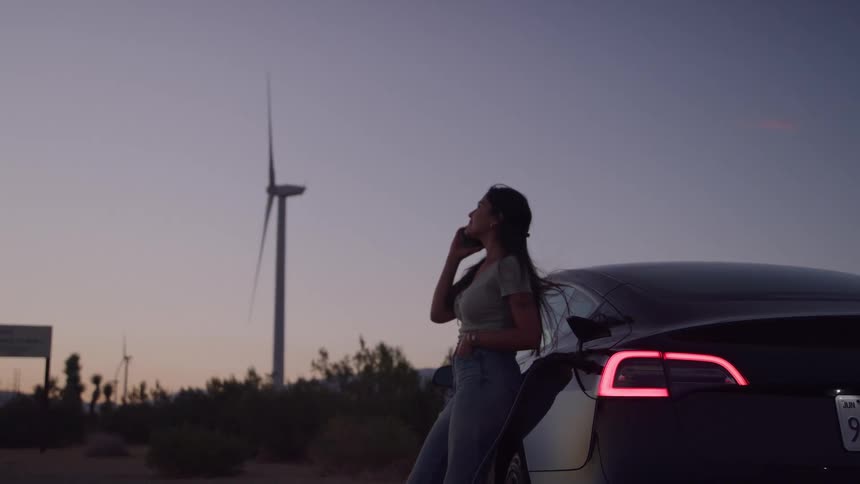 Woman leaning against a car while talking on the phone at sunset