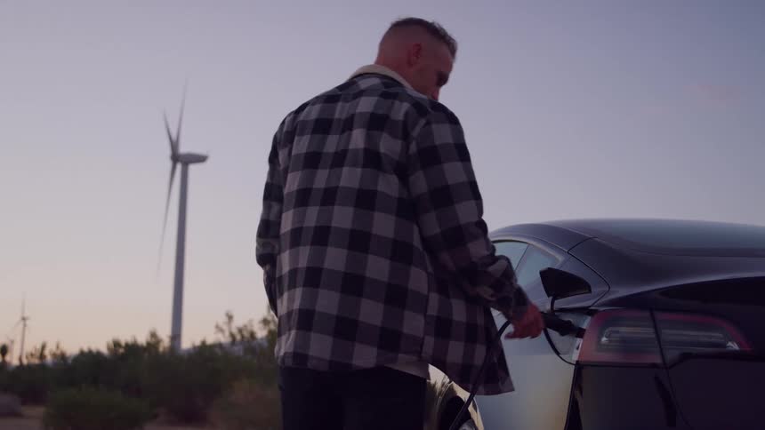 Man walks to parked car and plugs in an electric charger as wind turbines spin in the distance