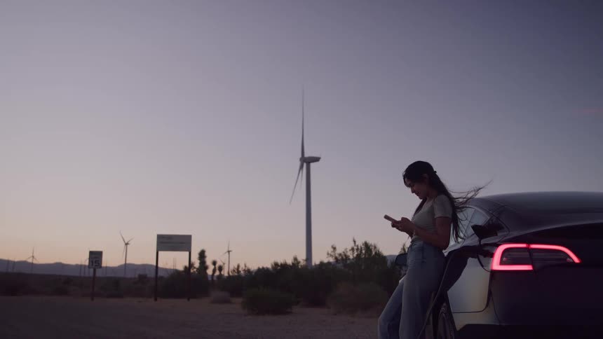 Woman leaning against a car looking at her phone at sunset near wind turbines
