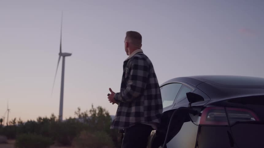 Man walks to parked car and plugs in an electric charger and leans against the car holding his phone as wind turbines spin in the distance