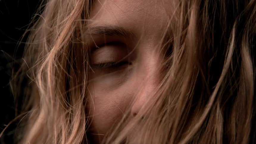 The video is an extreme closeup shot of a persons eye and the bridge of their nose framed by loose windblown blonde hairthe eye is partially obscured and slightly closed before opening fully to stare intently at the camera conveying a sense of em