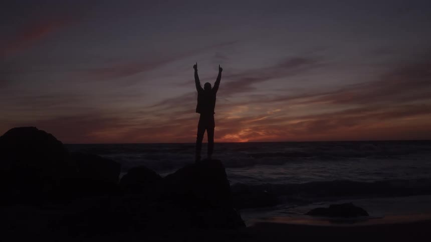 A man stands on a beach with his hands raised as the orange sunset sky glows behind him
