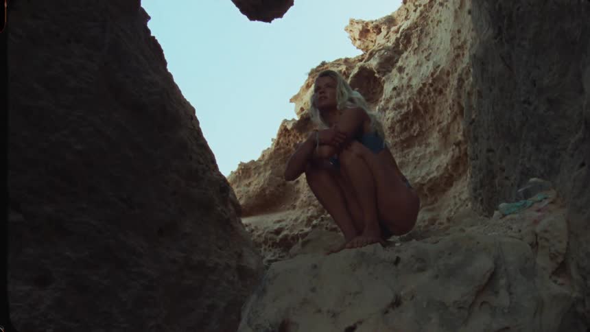 A beautiful woman with long blonde hair wearing a blue bikini is crouching on a sunlit rock ledge inside a dark cave or natural rock opening
