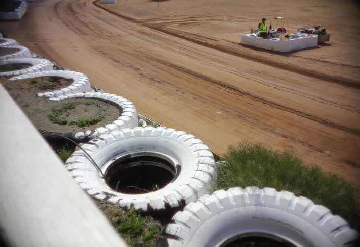 A dirt racetrack curves past a row of large white painted tire barriers with two workers at equipment in the distance