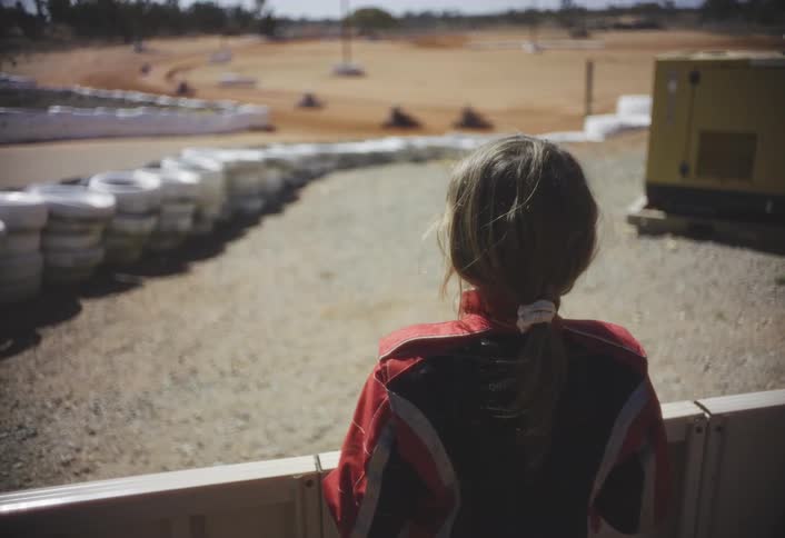 A girl far back from the track is watching motorcycles race