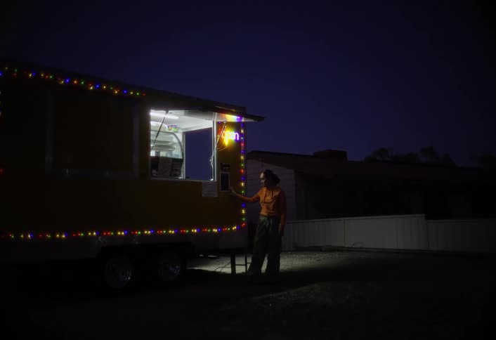 A person stands beside a brightly lit yellow food truck decorated with colorful string lights at night