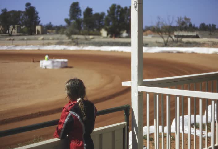 A young girl seen from the side and back stands leaning on a dark railing of a viewing platform intently watching a kart race quickly across a wide sunlit redbrown dirt track stretching out before her with a line of trees visible in the distance