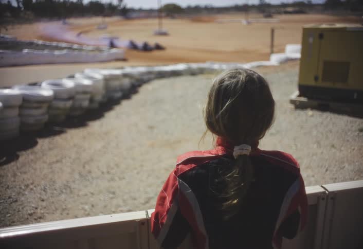 A person wearing a red and black racing jacket stands with their back to the camera looking out over a dirt racetrack the track is lined with stacked white tires and features a yellow generator in the background