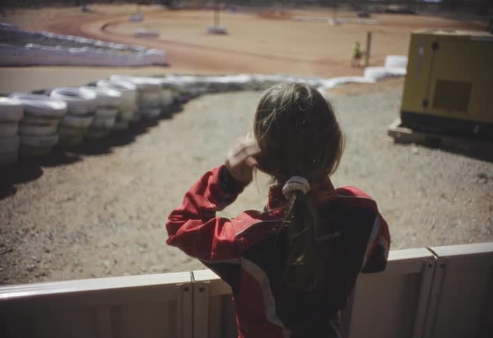 A young girl watches another person drive a go kart on a track