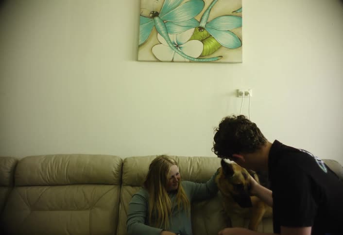 A brother and sister sit on a couch petting their dog in a cozy living room