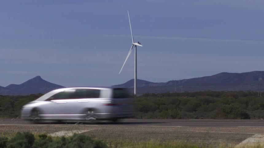 A wind turbine stands centered in the landscape the turbine blades turn slowly several cars pass the road mountains form the background and theres a blue sky with clouds