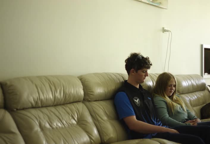 A teenage boy and a girl sit spaced apart on a beige leather couch in a plain living room looking bored and quiet