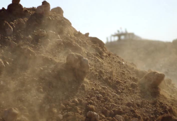 A yellow excavators bucket raises high and then drops a large pile of dark brown soil and rocks creating a cloud of dust under a bright sunny sky on a construction site