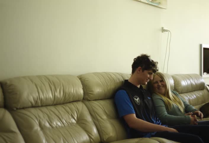 A young man and woman are seated closely together on a lightcolored leather couch in a bright room they initially smile and look at each other before turning their attention forward with matching relaxed smiles