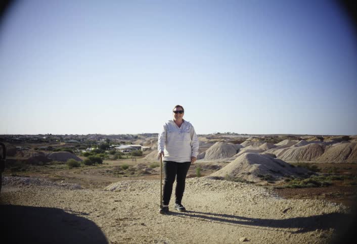 A person stands in a sunny arid landscape holding a walking stick with numerous piles of dirt or tailings behind them in the distance a small town or settlement is visible on the horizon beneath a clear blue sky
