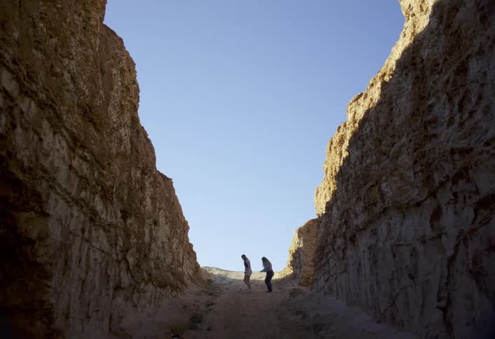A distant man and woman are dancing on a steep sandy path between tall light brown earthen canyon walls toward a bright blue sky opening