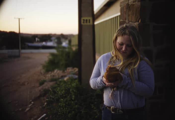 A teenager stands outside near a rustic building gently holding a chicken at dusk