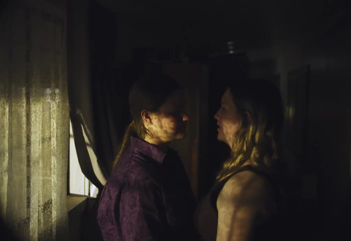 Two people stand facing each other in a dimly lit room with one person gently touching the others head the only significant light source comes from a window on the left which casts shadows across the figures