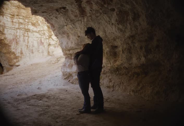 A couple is standing together in a dark natural cave or grotto embracing each otherthey are silhouetted against a large opening to the left which reveals a brightly lit tancolored wall outside or deeper within the cavern