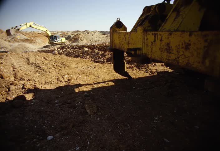 A close view of the large yellow frame of a piece of heavy equipment frames a sunny brown dirt construction site where a distant yellow excavator is digging a pit into a large pile of earth