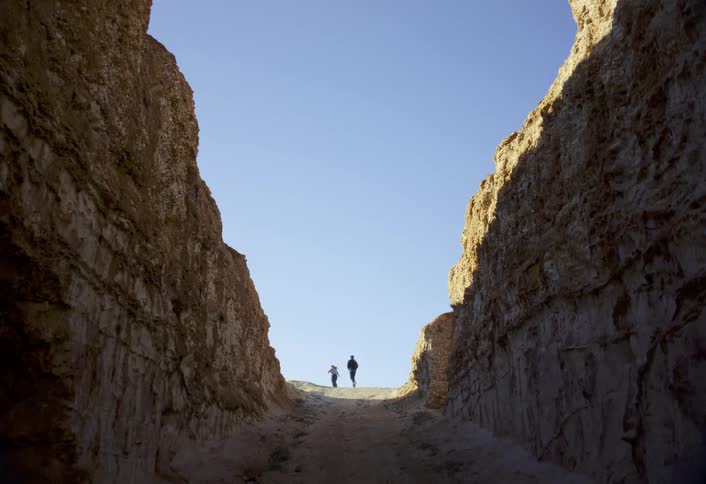 Two people run on the path between tall earthen walls during the daytime