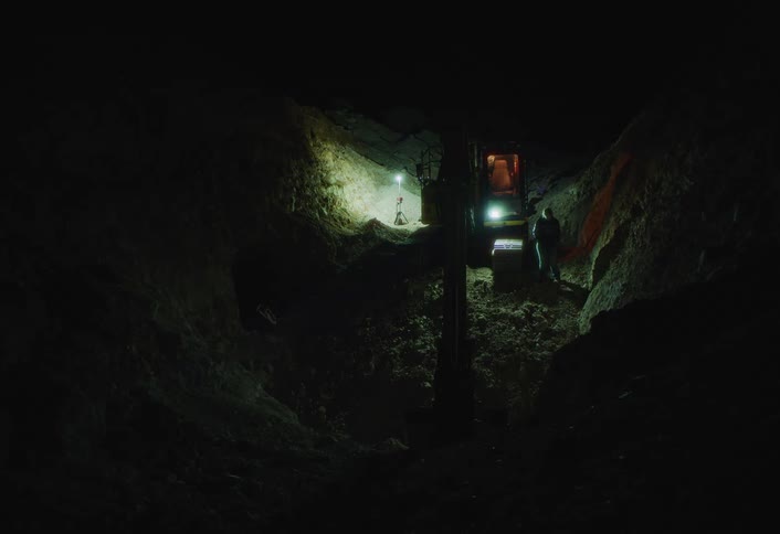 Workers operate an excavator at a nighttime dig site lit by floodlights