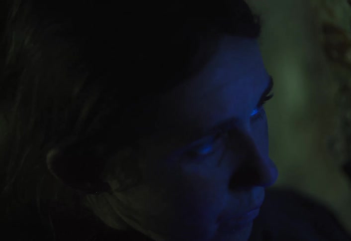 A dark close view shows a womans face illuminated by a flickering blue light source reflecting in her eyes creating a dramatic and mysterious atmosphere