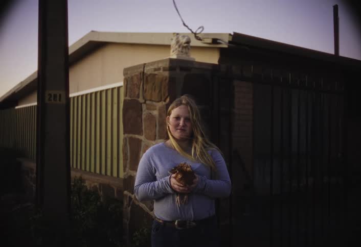 A woman stands holding a chicken in front of a house at dusk