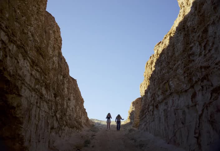 A deep rock ravine opens to a clear blue sky as two women walk and dance near the sunlit exit