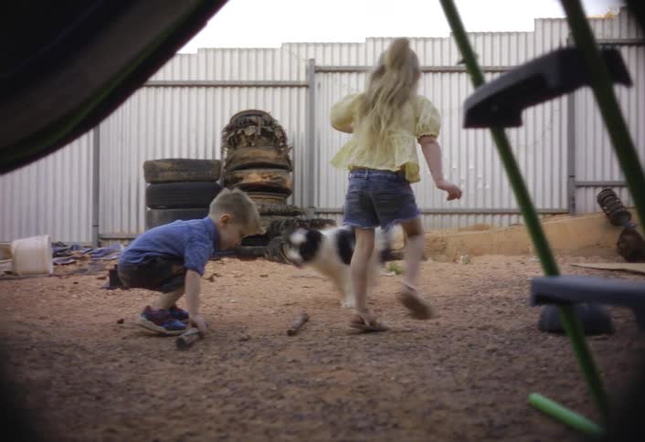 Two kids joyfully play with their dog in a dusty fenced backyard