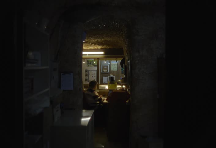 A dim wide interior view shows a cave like structure with two people sitting at a desk and working under yellow strip lighting at the end of a dark narrow corridor lined with rough earthen walls