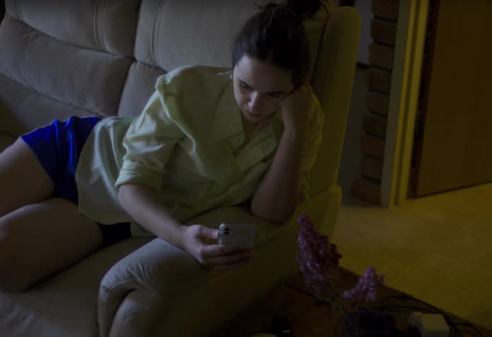 A woman lies on a couch talking on a video call in a dimly lit room