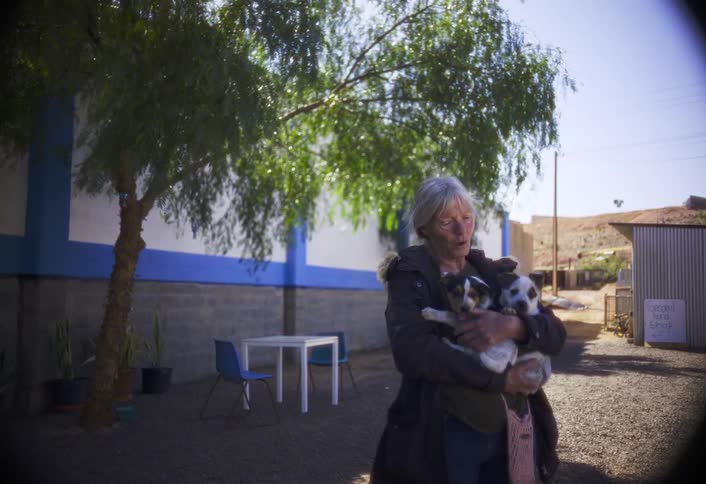 A smiling older woman with grey hair stands under a tree in the bright sunlight talking happily as she holds two small spotted puppies in her arms