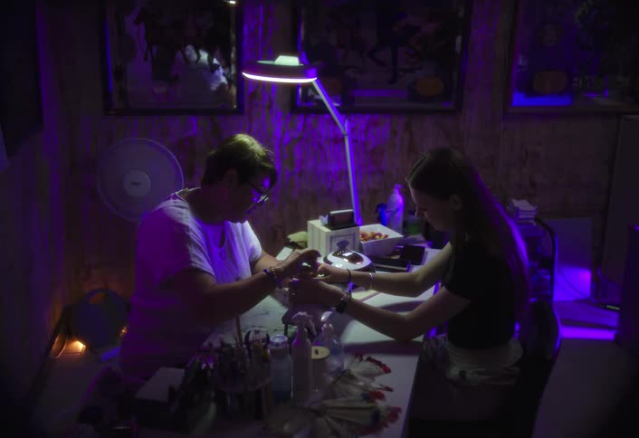 A nail artist works on a clients hands under a lamp in a dimly lit room