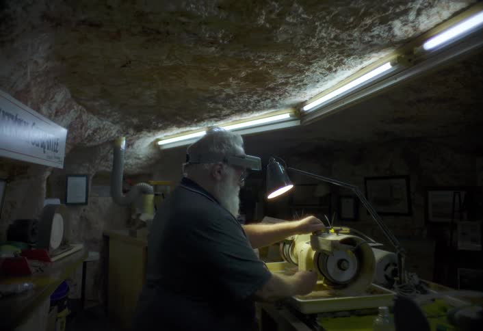 An older man works on a machine under a lamp in a dim underground workshop